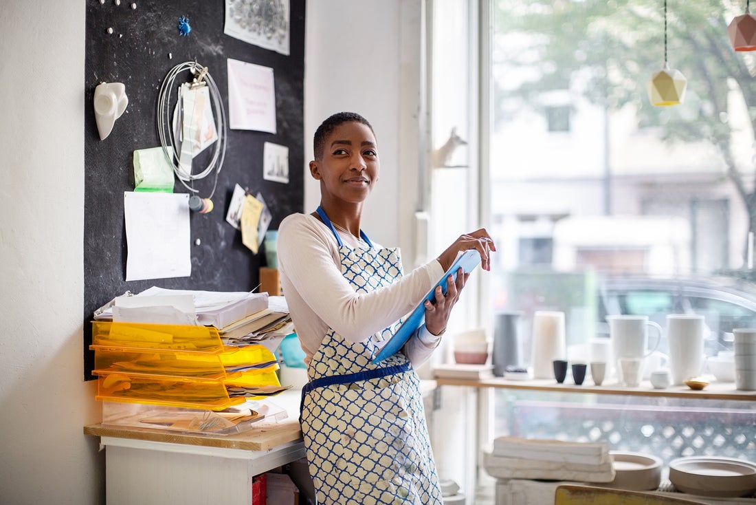 A young woman stands in a pottery studio, smiling while holding a blue tool. Bright natural light streams through the large window, showcasing various ceramics on display.