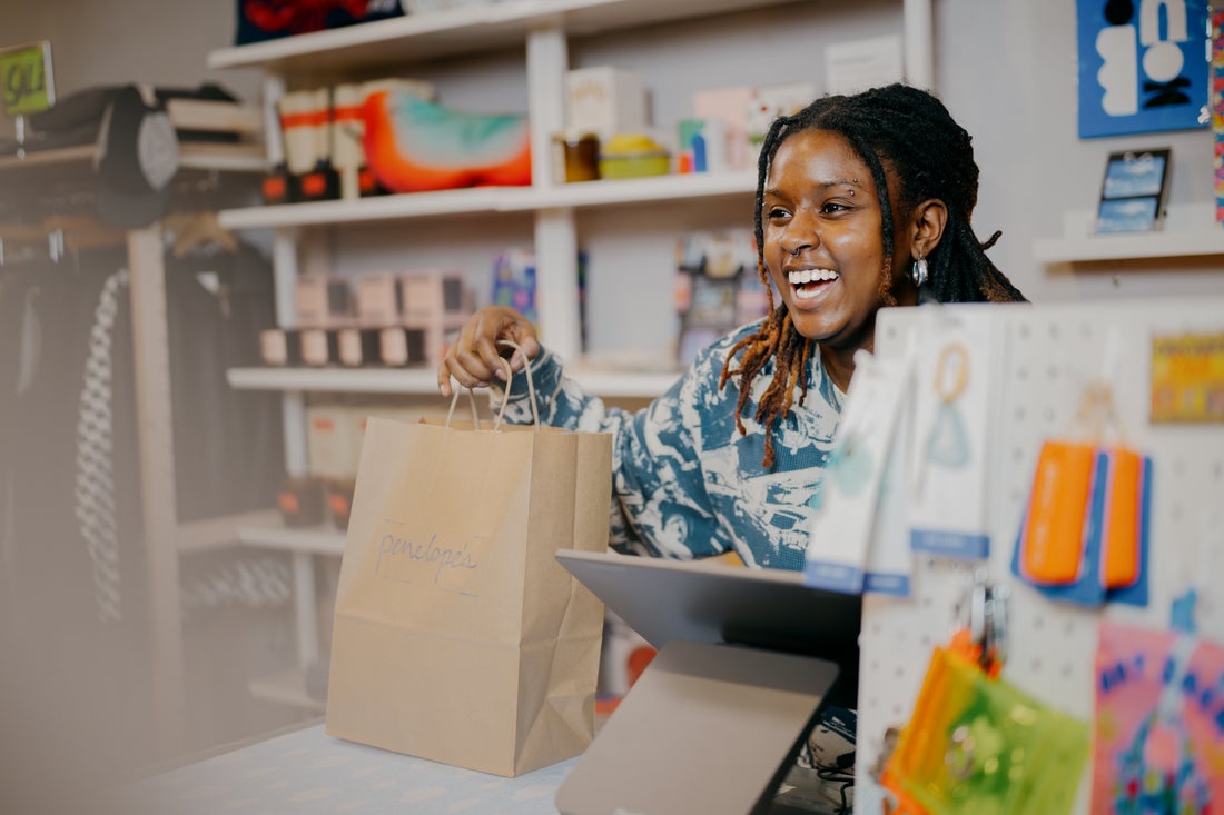 A smiling cashier holds a shopping bag labeled "penelope's" at a colorful retail store, showcasing a friendly shopping experience.