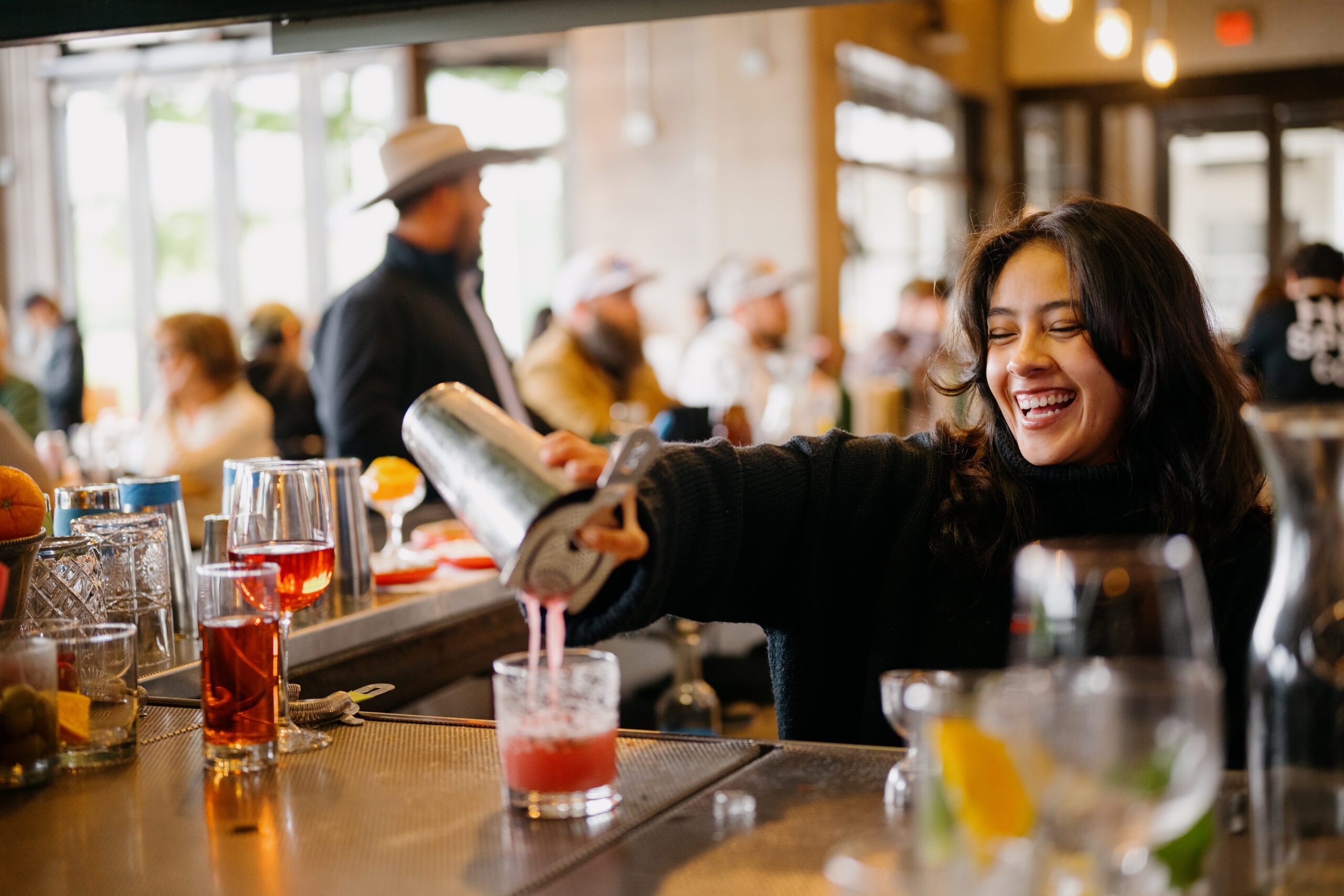 A bartender joyfully pours a pink cocktail from a shaker into a glass, showcasing a vibrant bar atmosphere filled with patrons.