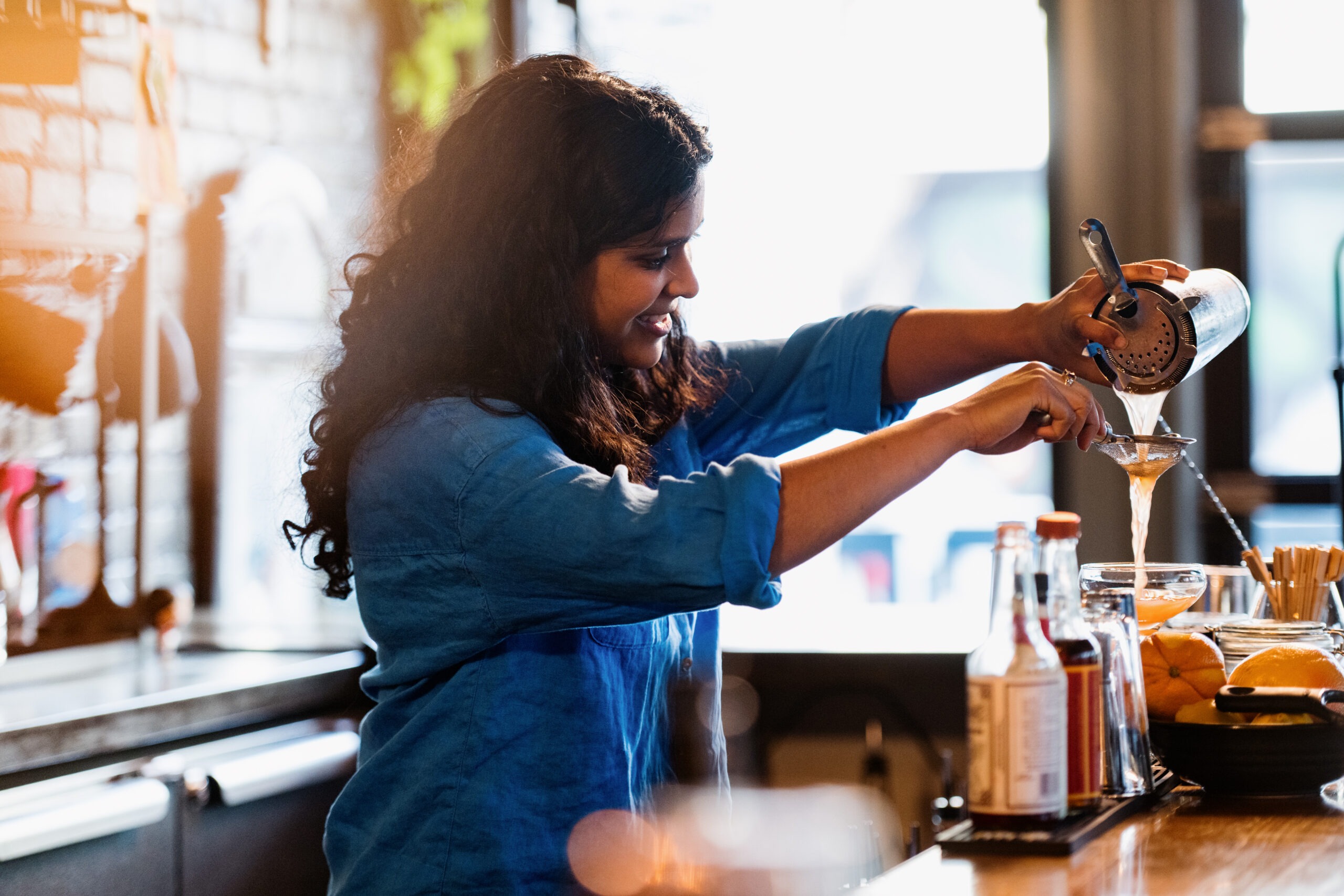 A bartender strains a cocktail into a glass, showcasing skill and attention to detail. Bottles and fresh ingredients are visible on the counter.