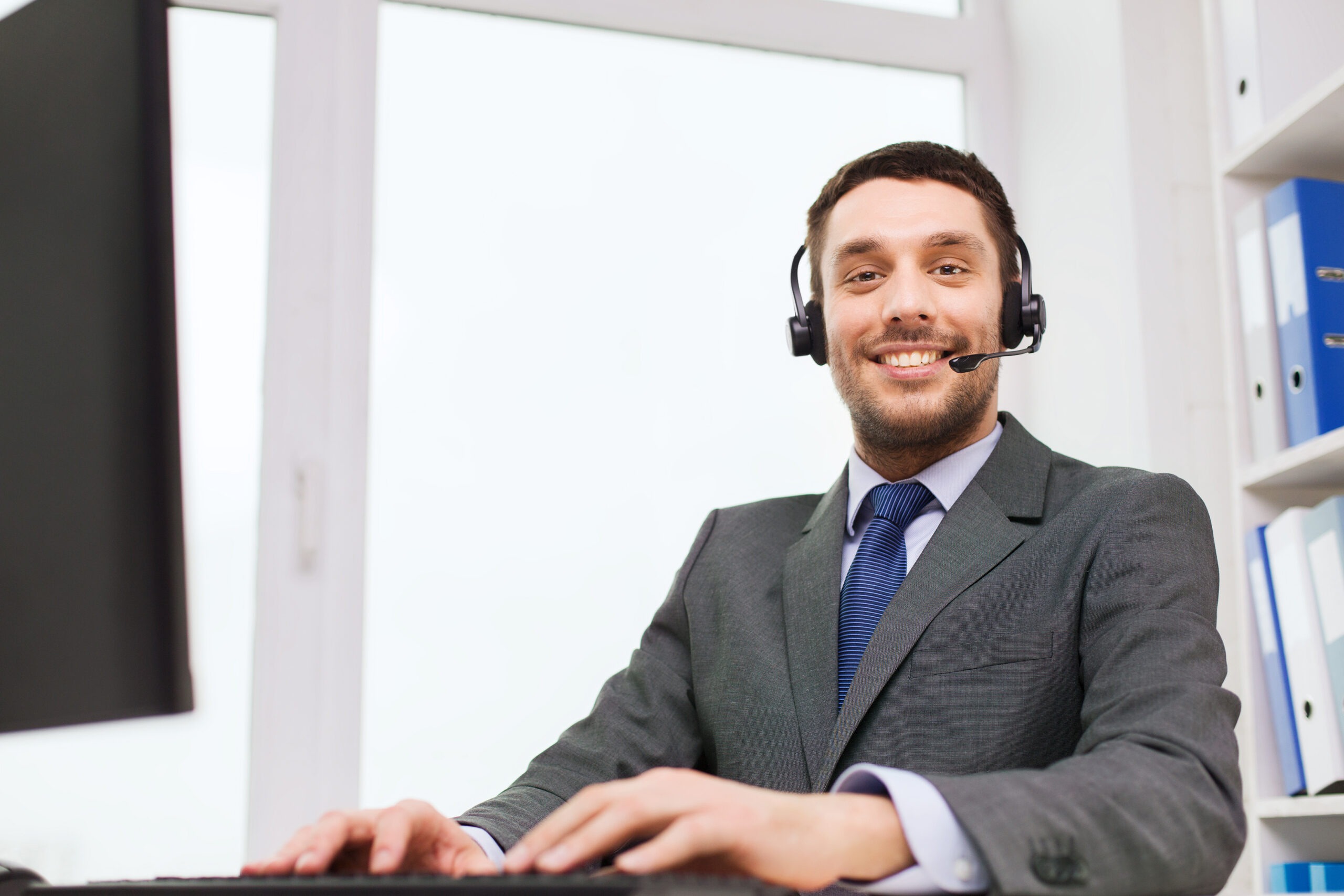 Professional man in a suit and headset smiles at the camera while working at a computer, representing customer service or remote work.