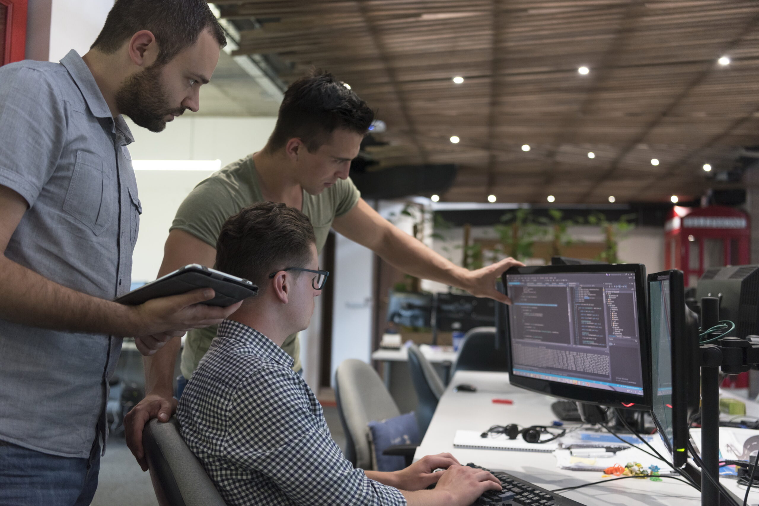 Three software developers collaborate at a modern workspace, reviewing code on a monitor. One holds a tablet while another explains details.