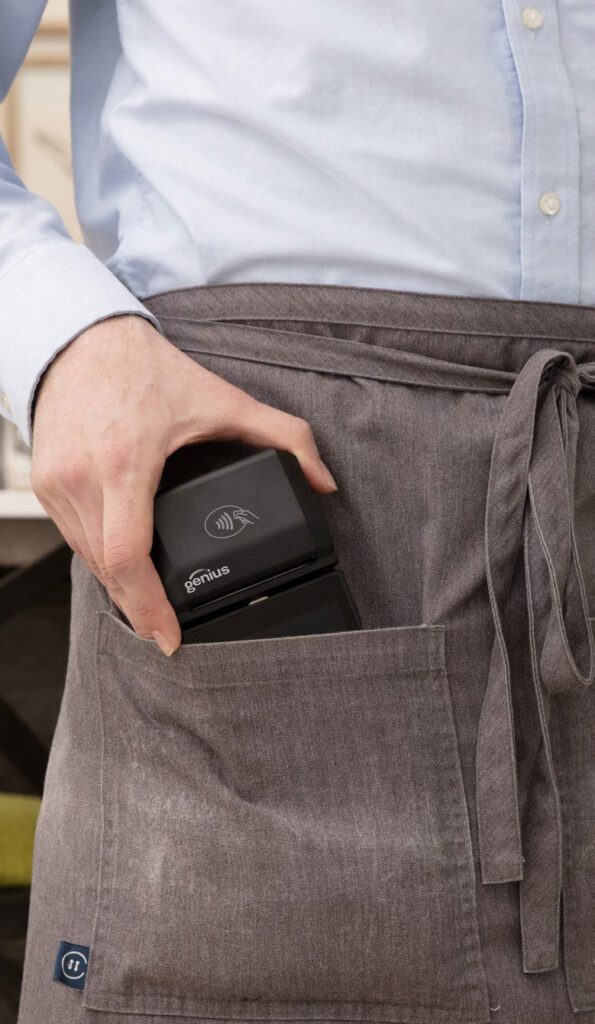 A person holds a black portable kitchen device labeled "genius" as they place it into the pocket of a gray apron, indicating convenience in cooking.