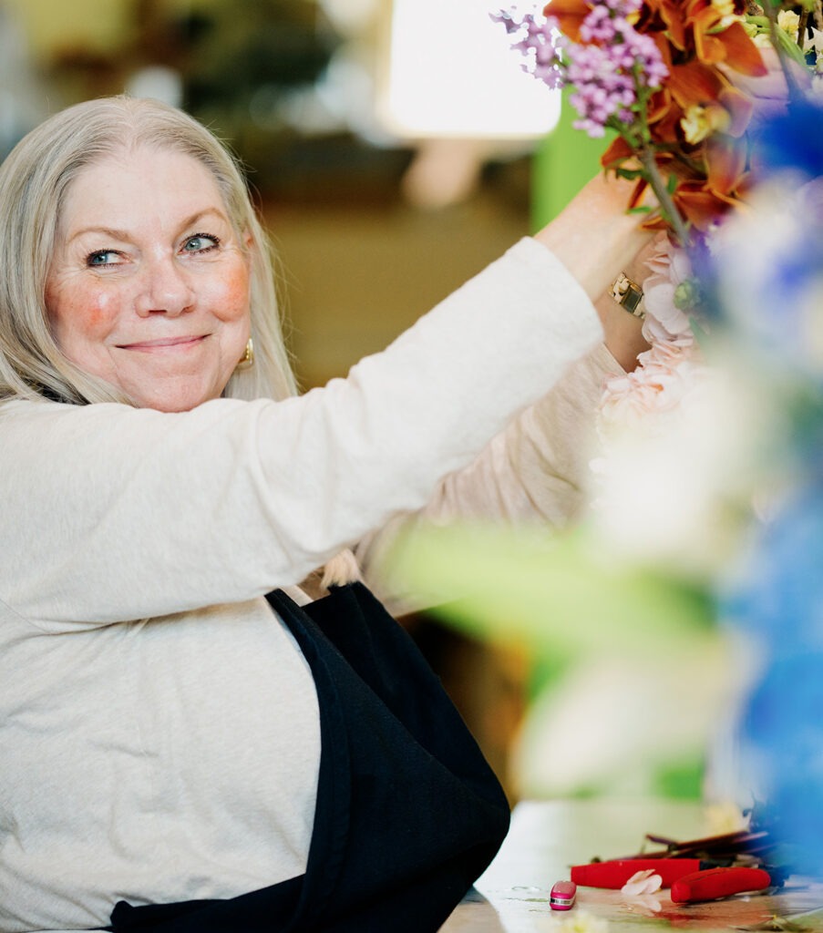 Florist arranging colorful flowers, smiling warmly. The vibrant setting highlights her passion for floral design, enhancing the shop's inviting atmosphere.