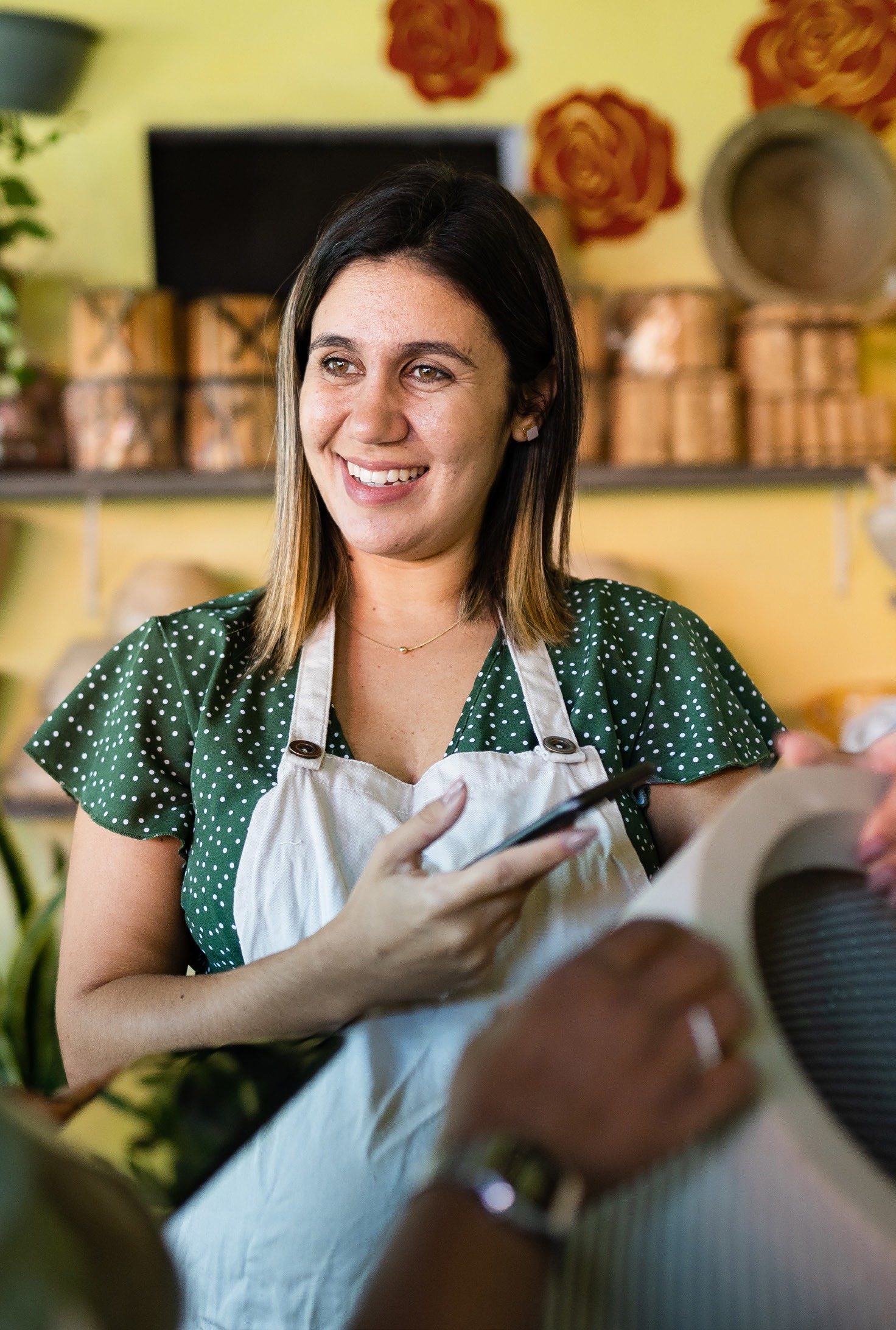 Smiling woman in an apron holds a phone, engaging with a customer in a vibrant cafe adorned with plants and decorative items.