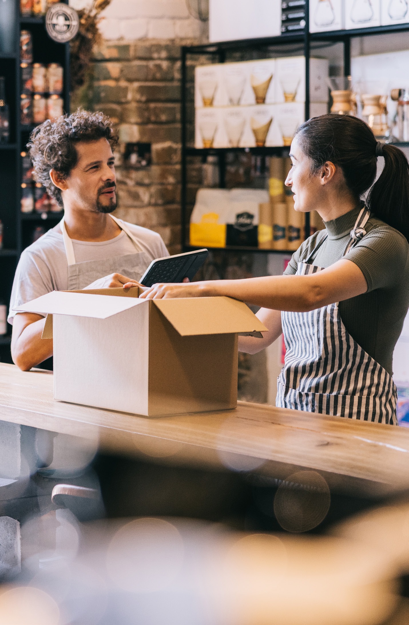 A customer interacts with a barista at a café, discussing items inside an open box on the counter while using a tablet, emphasizing customer service.